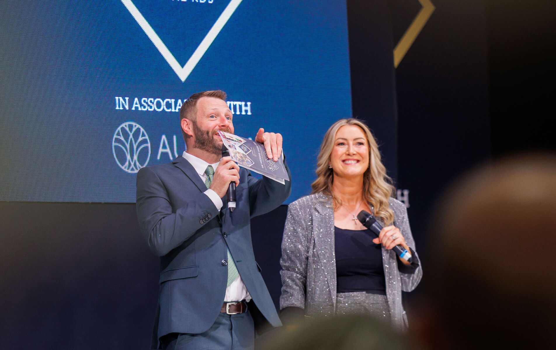 Two hosts on stage presenting at an awards event, both smiling and holding microphones.