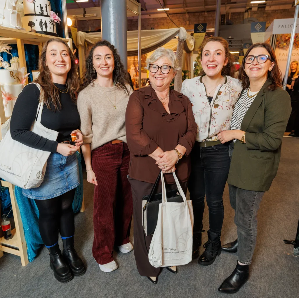 Group of smiling women at an indoor event, holding bags, standing together, enjoying a social gathering.