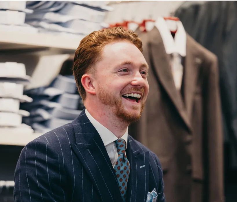 Man in a suit smiling in a clothing store with racks of shirts and jackets in the background.