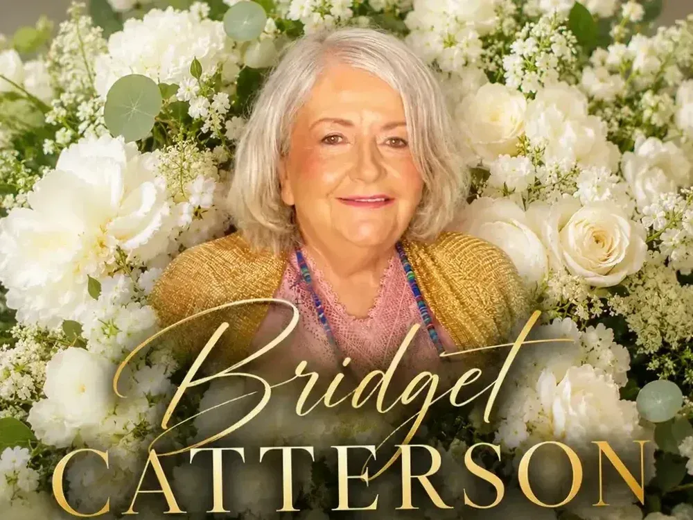Elderly woman smiling in front of a floral backdrop, wearing a colorful outfit with text Bridget Catterson.