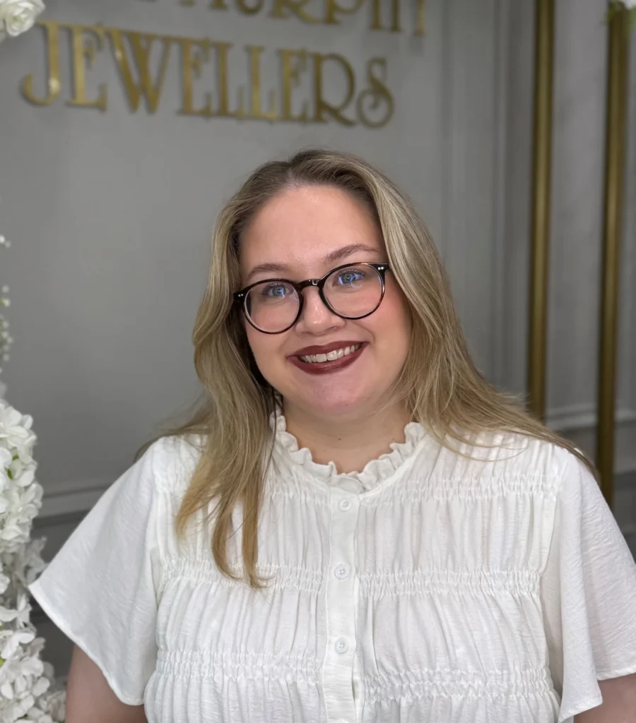 Smiling woman in glasses with a white blouse, standing in front of a jeweller's sign.