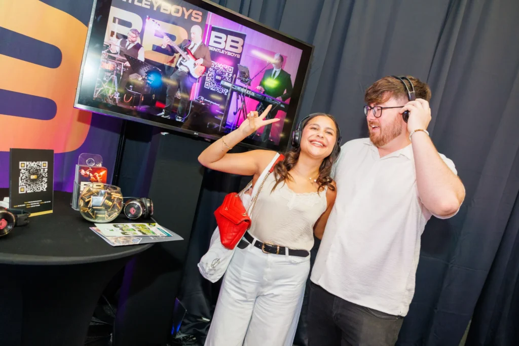 People enjoying music at an event booth with a live band displayed on a screen in the background.