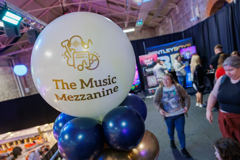 Balloon with The Music Mezzanine logo at a lively event with people and colorful displays in the background.