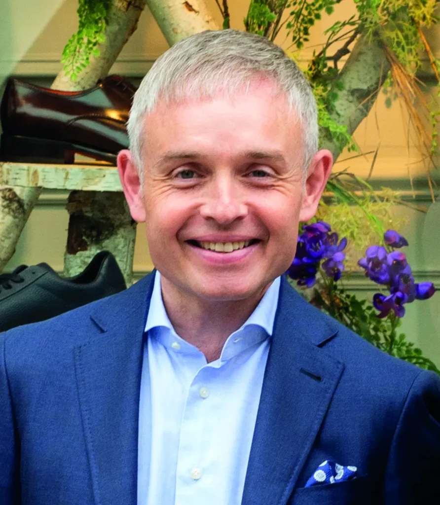 Smiling man in a blue suit in front of a natural background with shoes and flowers.