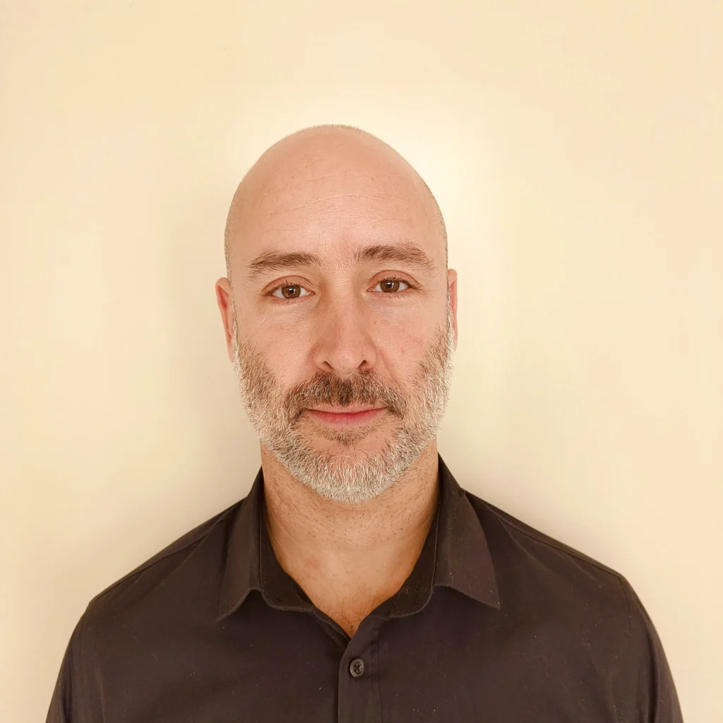 Bald man with a beard in a black shirt against a beige background, looking at the camera.