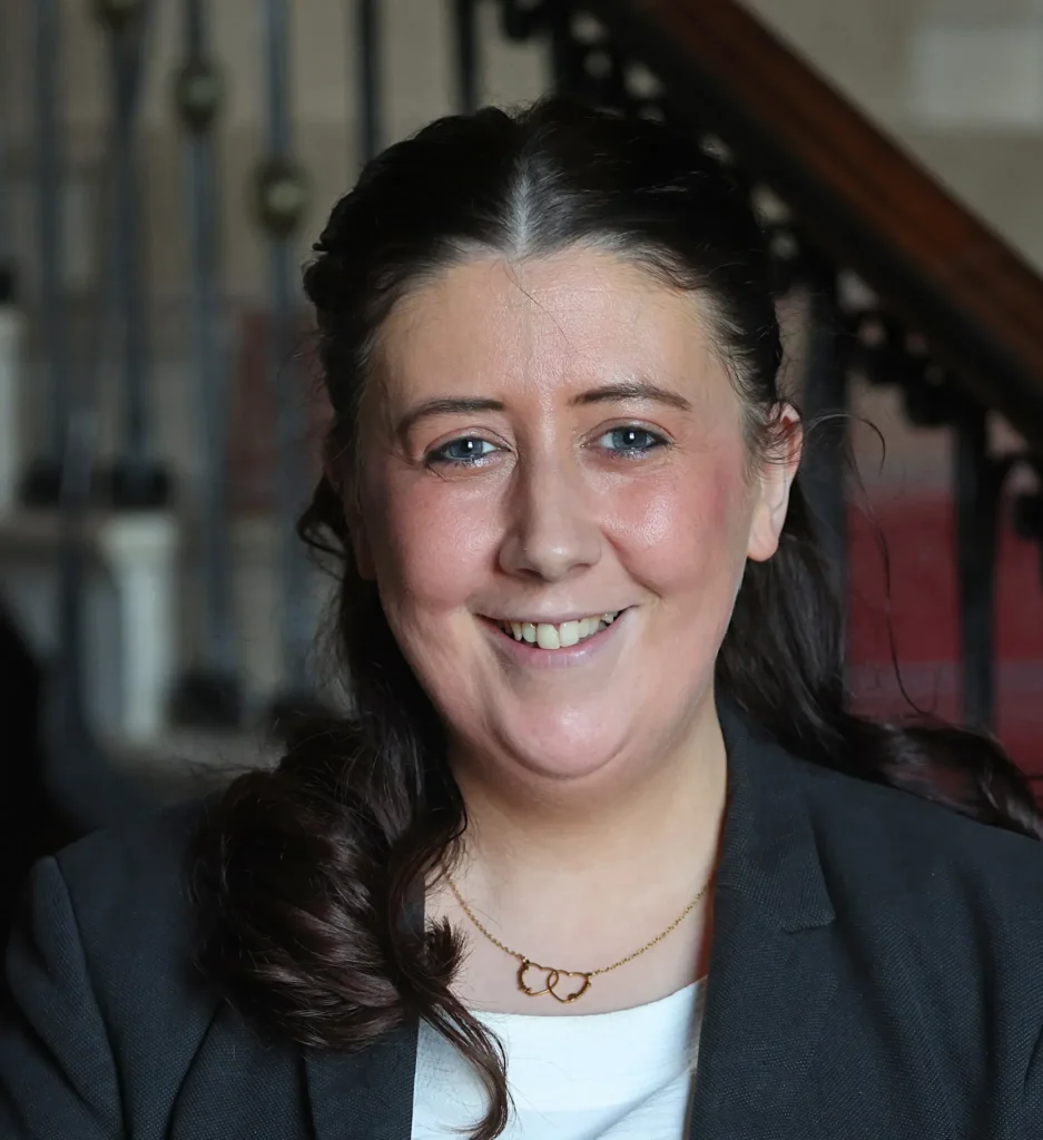A person smiling indoors, wearing a blazer and heart necklace, with a blurred staircase in the background.