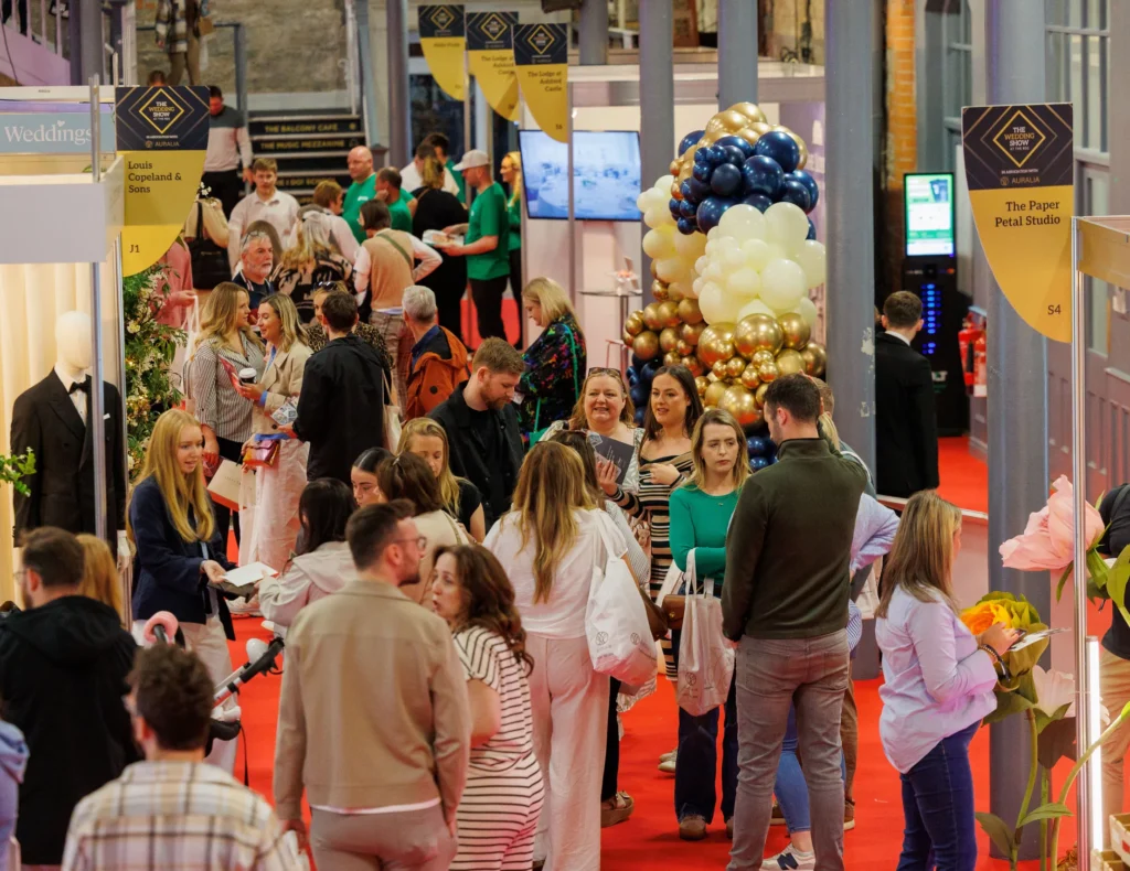 Busy wedding expo with attendees engaging at booths, vibrant balloon decor, and red carpet ambiance.