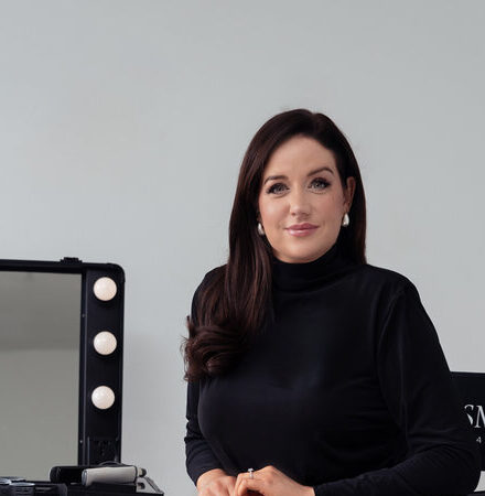 Woman in black top seated at a makeup station with lights and mirror, smiling confidently towards the camera.