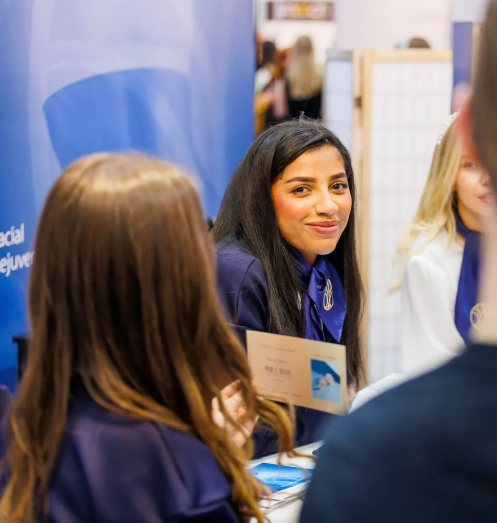 A smiling woman at a promotional event, engaging with attendees, holding a flyer against a blue backdrop.