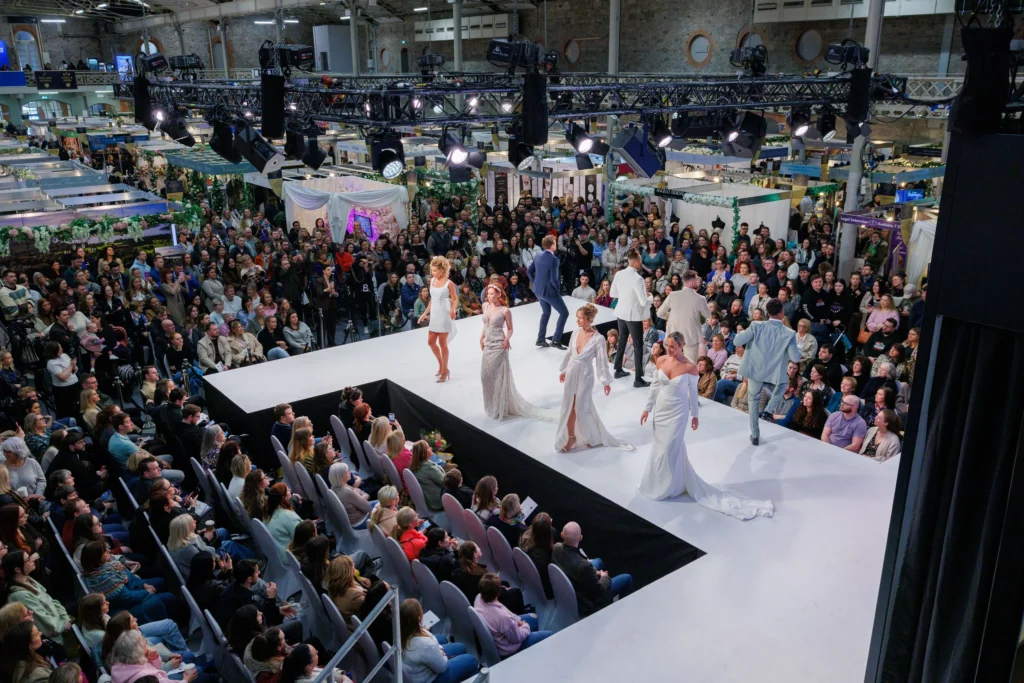 Fashion show with models in elegant attire on stage, surrounded by audience at a crowded event.