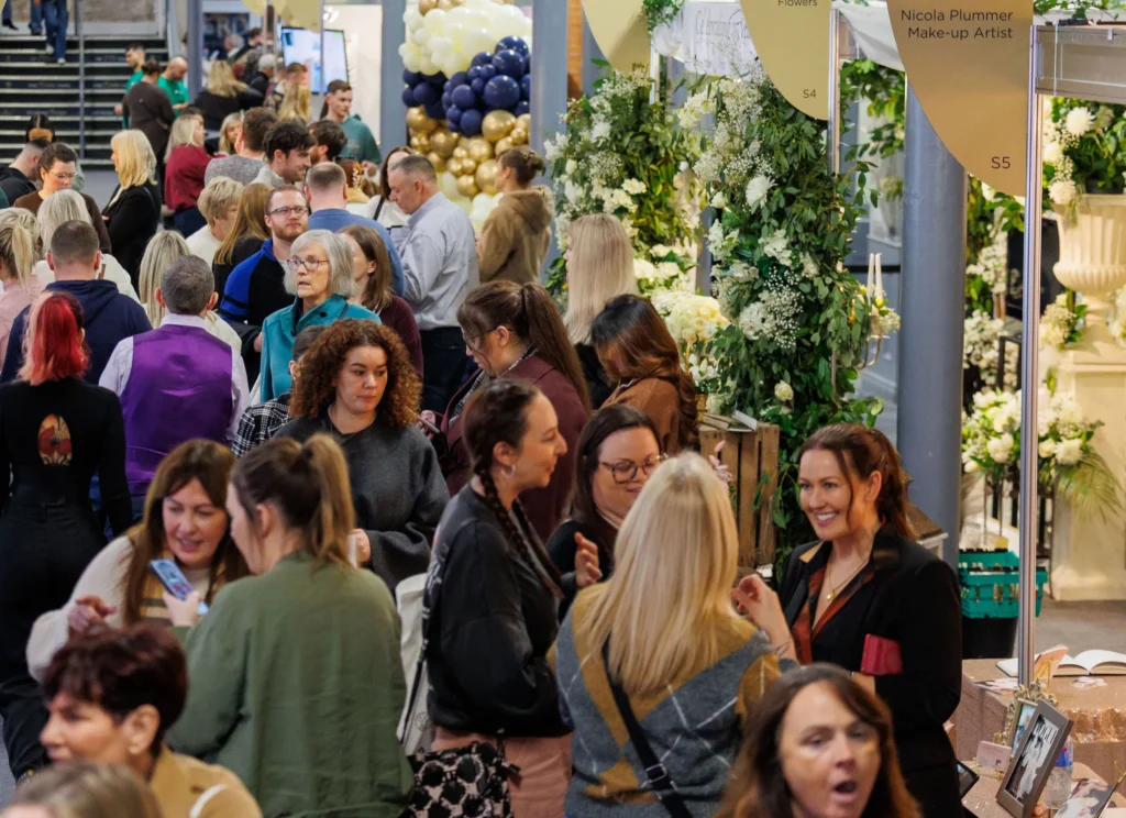 Crowded wedding exhibition with attendees exploring floral and makeup stands amidst decor and vibrant atmosphere.