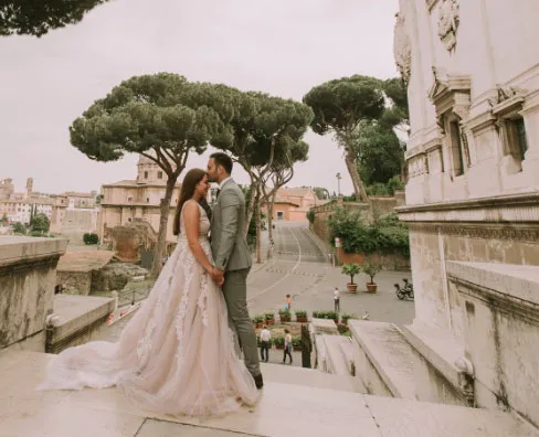 Bride and groom share a kiss on historic stone steps, surrounded by scenic architecture and lush trees.
