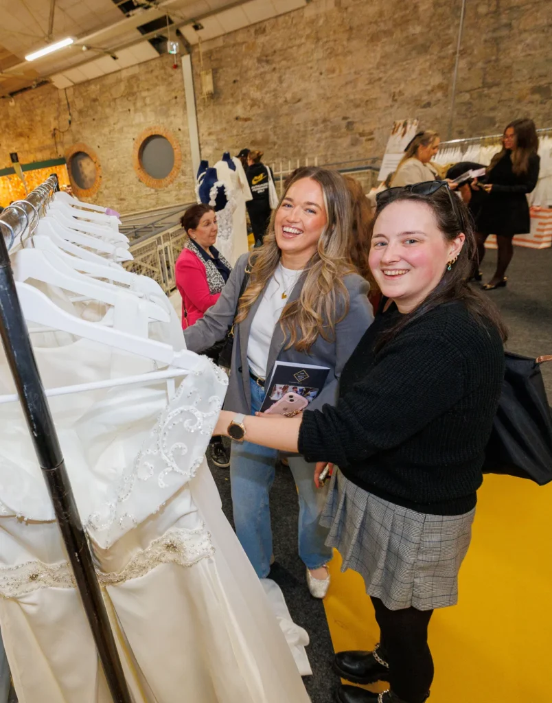 Two women smiling and browsing wedding dresses at a bridal store event with elegant white gowns on display.