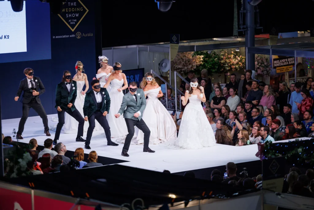 Models in wedding attire dance on stage at a bridal show with audience watching eagerly.