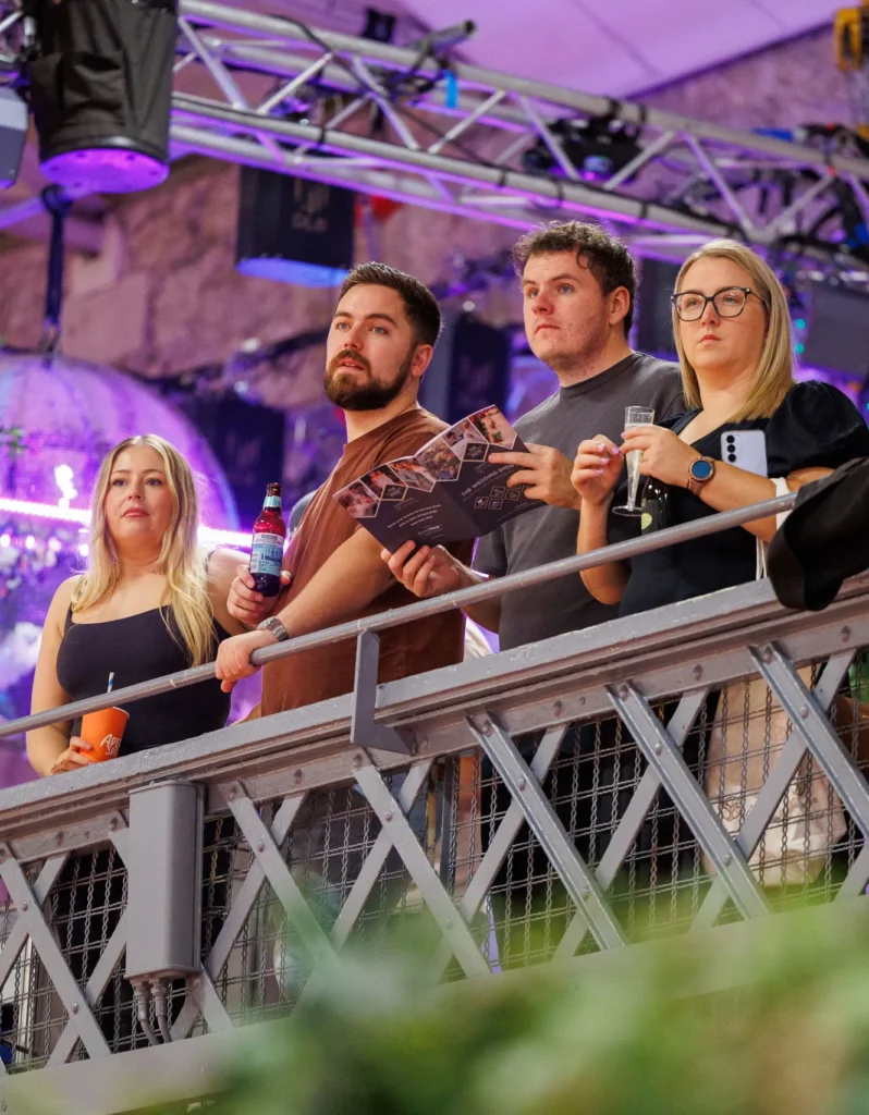 A group of people attentively watching an event from a balcony with colorful lighting overhead.