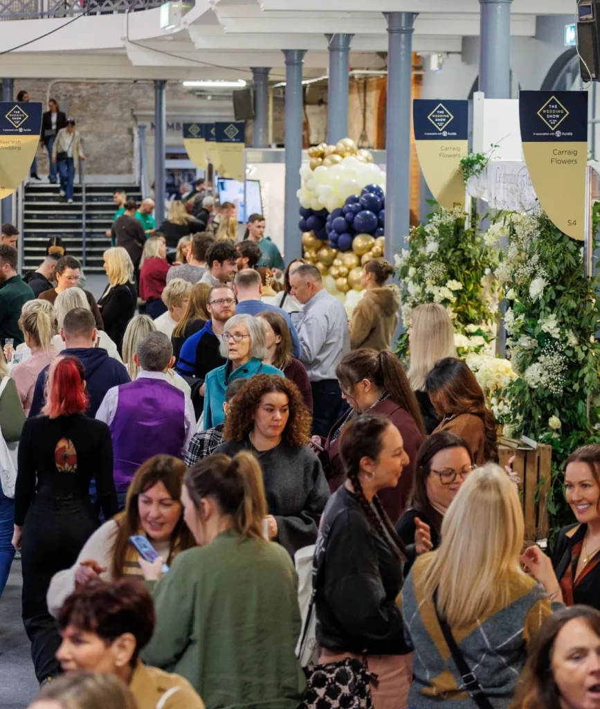 Crowds attending a lively exhibition with floral displays and balloons, highlighting vendor booths and social interactions.