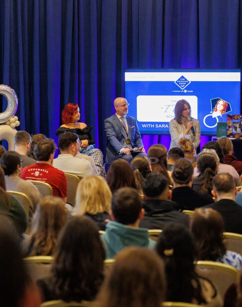 Panel discussion at a conference with audience, featuring speakers and a backdrop screen displaying event details.
