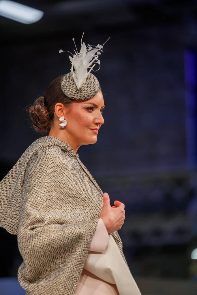 Fashion model in elegant ensemble with stylish fascinator and textured cape, captured during a runway show.