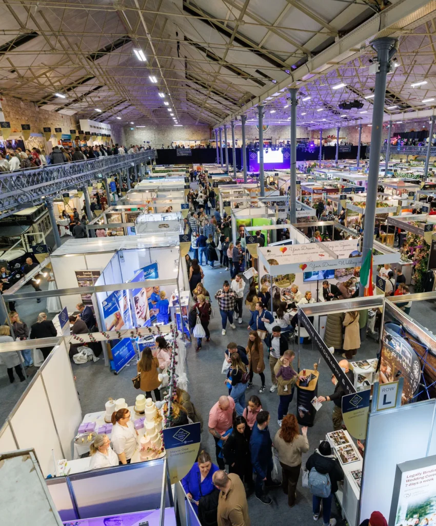 Busy trade fair with people exploring various booths inside a large, well-lit exhibition hall.