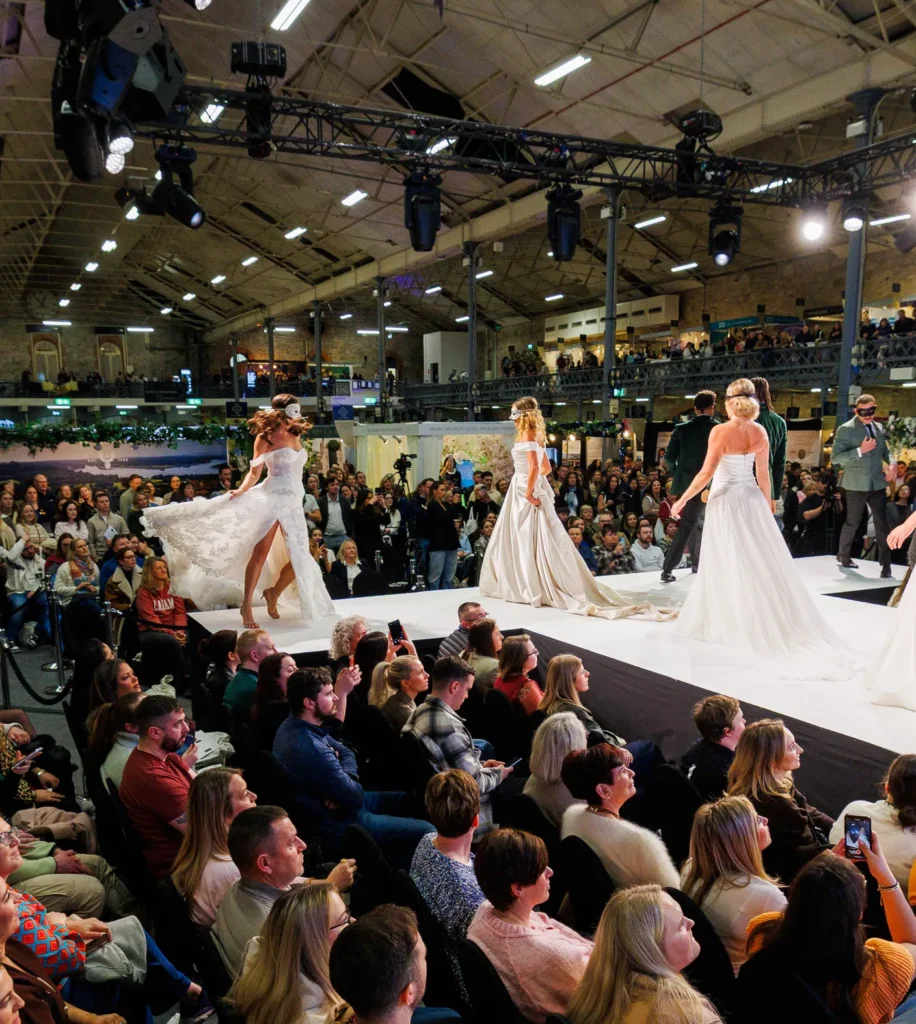 Bridal fashion show on runway with models in wedding dresses, engaging audience in indoor event space.