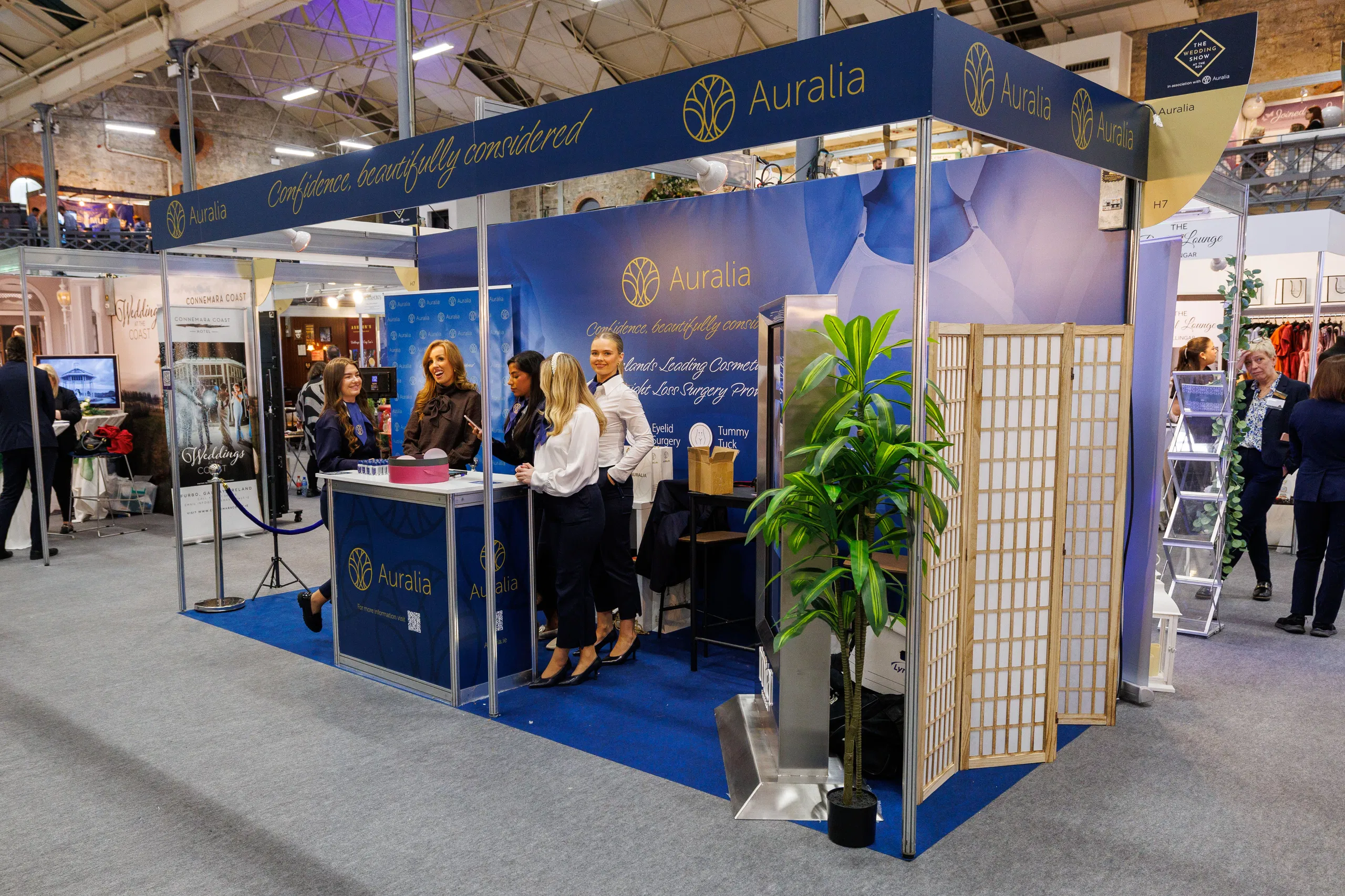 Auralia exhibition booth at a trade show with staff engaging visitors, showcasing cosmetic surgery services.