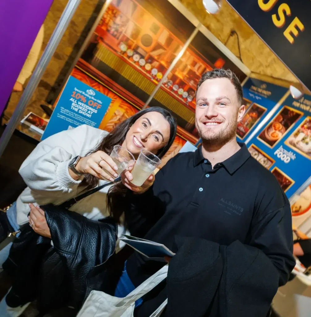 Smiling couple enjoying drinks at a festive event booth, surrounded by colorful posters and warm lighting.