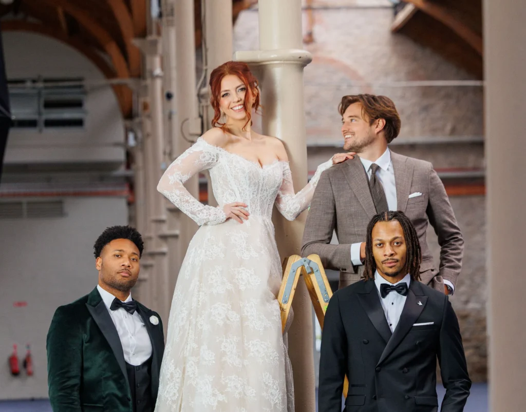 Bride in lace gown and three groomsmen in suits pose indoors with ladder and columns.
