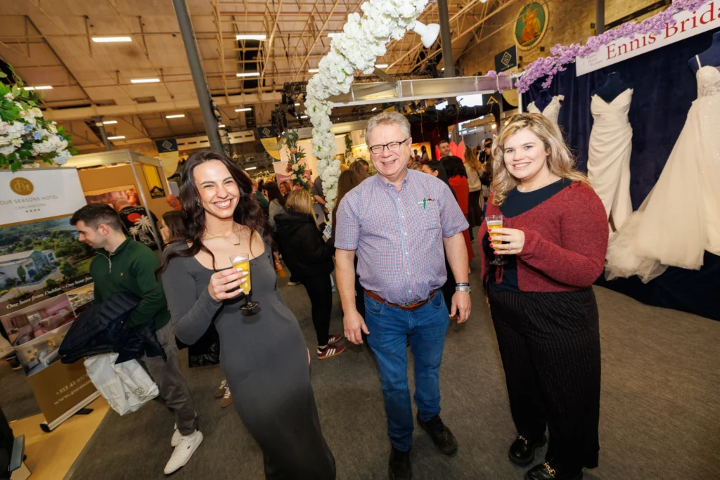 People enjoying drinks at a wedding expo, with bridal gowns and hotel brochures on display.