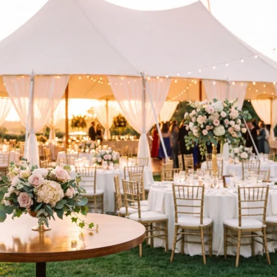 Elegant outdoor wedding reception with floral centerpieces, a white tent, and string lights at sunset.