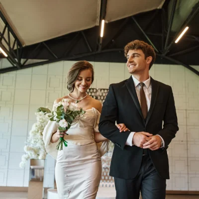 Happy couple walking arm-in-arm indoors, woman holding a bouquet, both dressed formally and smiling.