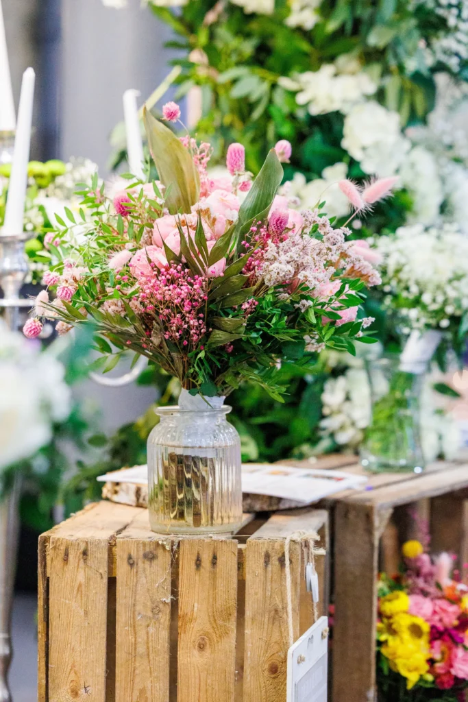 Pink floral arrangement in a glass vase on a wooden crate, surrounded by lush greenery.