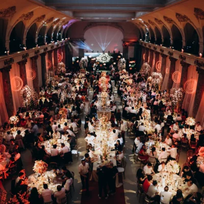 Elegant wedding reception in a grand hall with guests seated at decorated tables under warm lighting.
