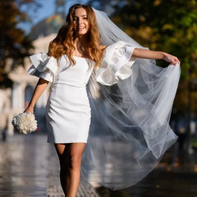 Bride in short white dress and veil, holding bouquet, smiles outdoors on sunny day.