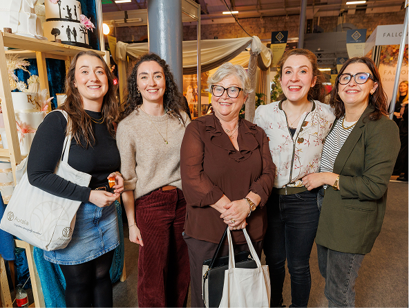 Group of five smiling women at a craft fair, standing together with bags and cakes in the background.