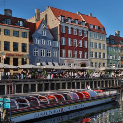 Colorful buildings and canal tour boat at Nyhavn, Copenhagen, on a sunny day, with people dining outdoors.