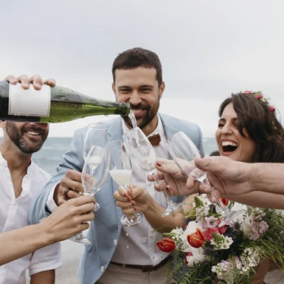 Group celebrating a beach wedding with champagne toasts, laughter, and a floral bouquet.